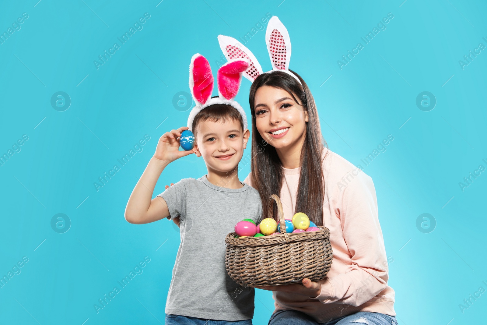 Photo of Mother and son in bunny ears headbands with basket of Easter eggs on color background