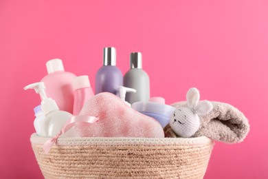 Photo of Different baby cosmetic products, bathing accessories and toy in wicker basket on pink background, closeup