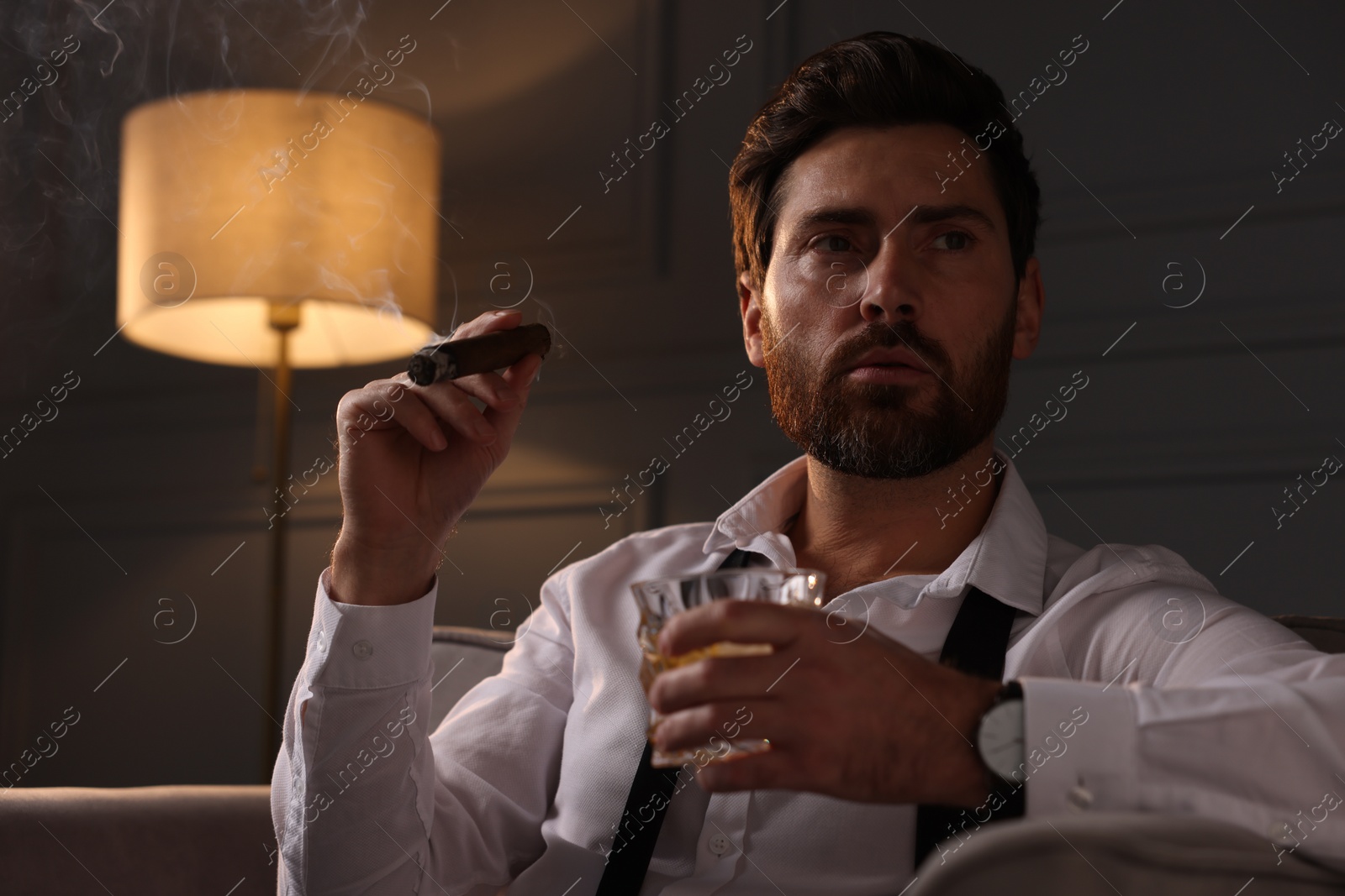 Photo of Handsome man with glass of whiskey smoking cigar at home