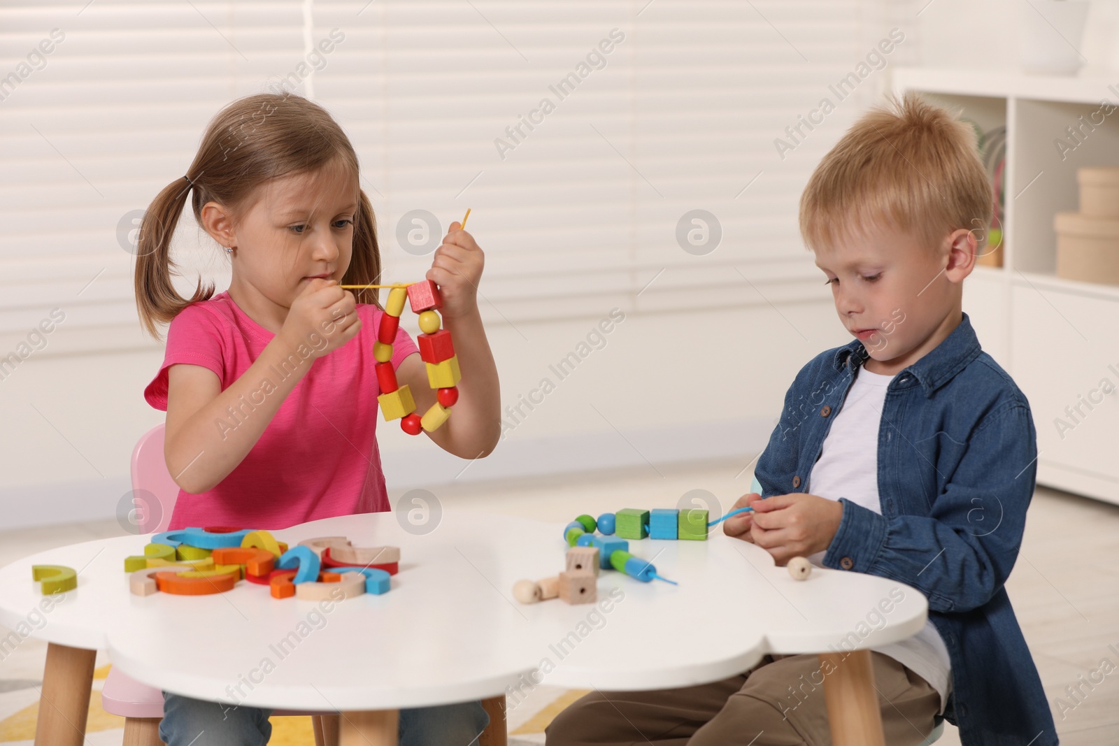 Photo of Little children playing with wooden pieces and string for threading activity at white table indoors. Developmental toys