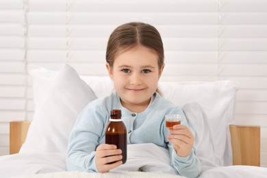 Cute girl holding bottle and measuring cup with cough syrup in bed. Effective medicine