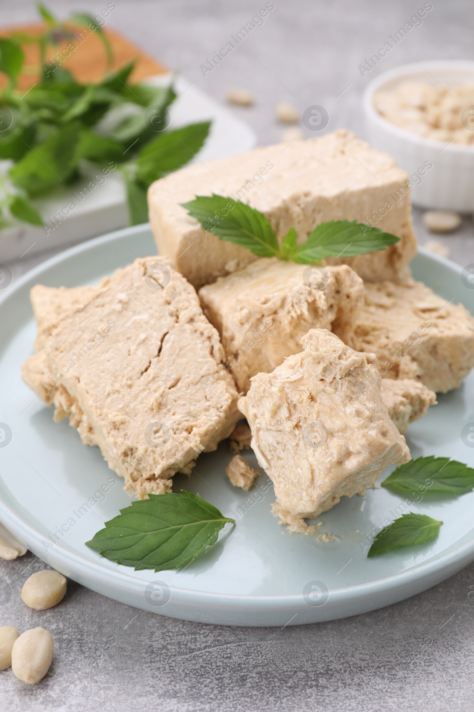 Photo of Plate with pieces of tasty halva, peanuts and mint leaves on light grey table, closeup