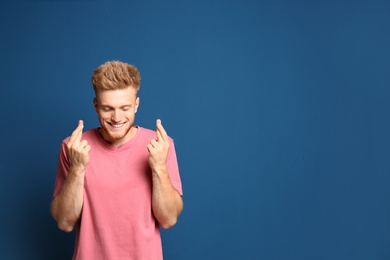 Portrait of hopeful man with crossed fingers on blue background, space for text