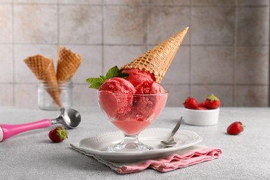 Delicious scoops of strawberry ice cream with mint and wafer cone in glass dessert bowl served on grey table