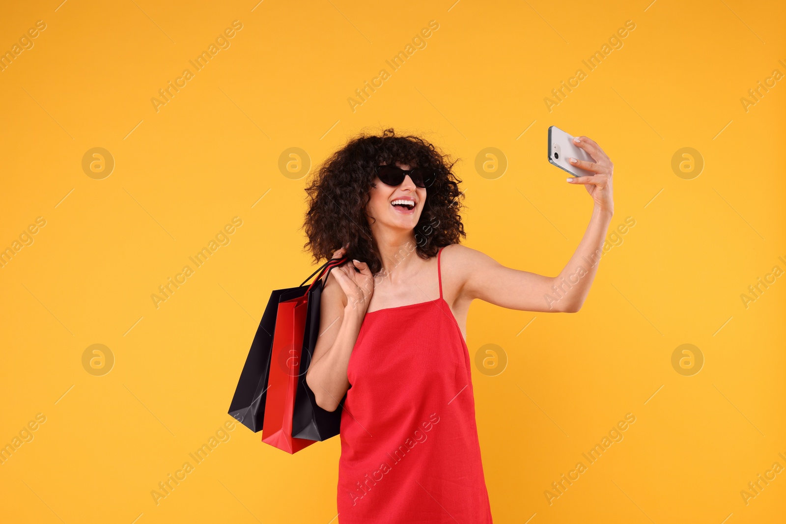 Photo of Happy young woman with shopping bags and stylish sunglasses taking selfie on yellow background