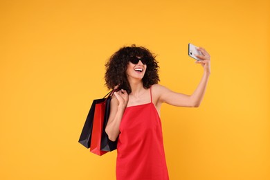 Happy young woman with shopping bags and stylish sunglasses taking selfie on yellow background