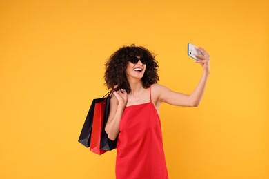 Photo of Happy young woman with shopping bags and stylish sunglasses taking selfie on yellow background