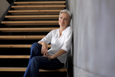 Photo of Happy mature woman sitting on wooden stairs indoors. Smart aging