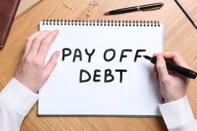 Image of Woman writing phrase Pay Off Debt in notebook at wooden table, top view