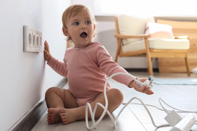 Photo of Cute baby playing with electrical socket and plug at home. Dangerous situation
