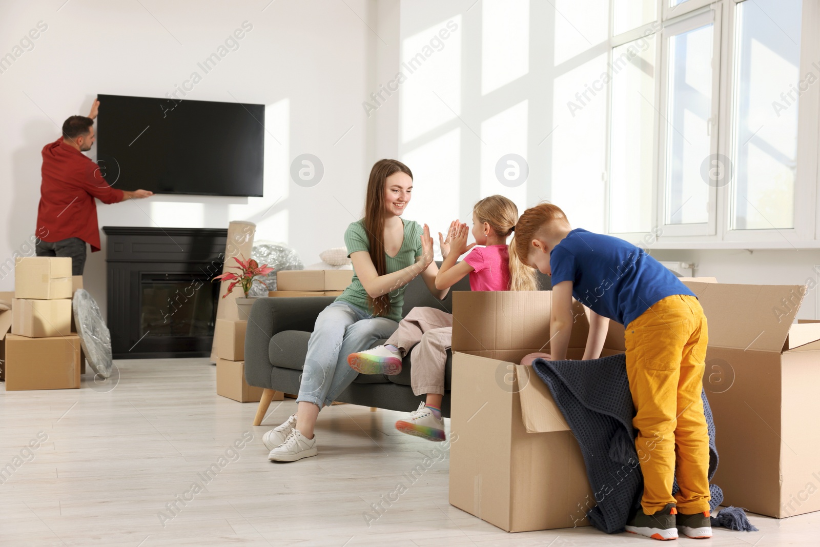 Photo of Family settling into new home. Dad setting up TV, happy mom and daughter playing on couch, son unpacking his boxes. Moving day