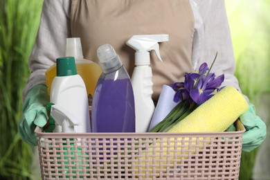 Photo of Woman holding basket with spring flowers and cleaning supplies outdoors, closeup