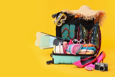 Open suitcase with clothes, beach accessories and shoes on yellow background, space for text. Summer vacation