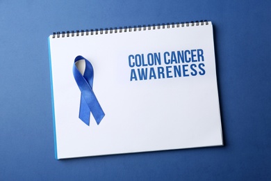 Photo of Notebook with blue ribbon and text Colon cancer awareness on color background, top view