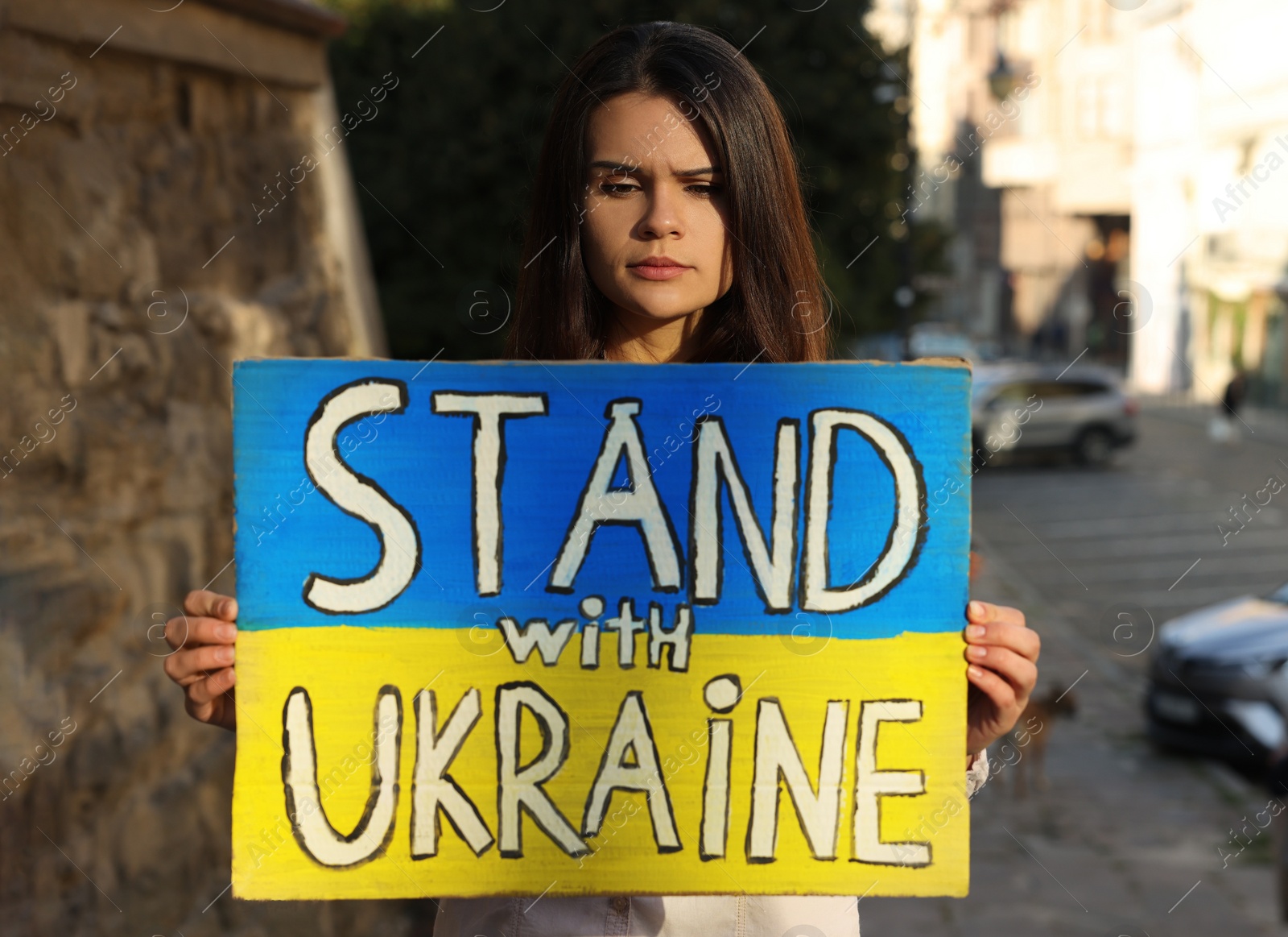 Photo of Sad woman holding poster in colors of national flag and words Stand with Ukraine on city street. Space for text