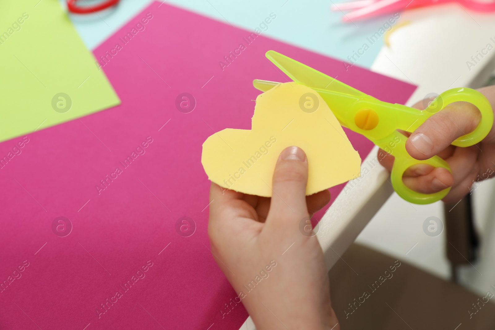 Photo of Child cutting out paper heart with plastic scissors at table, closeup. Space for text