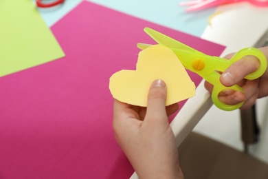 Photo of Child cutting out paper heart with plastic scissors at table, closeup. Space for text