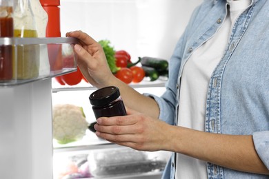 Photo of Young woman taking jar of jam out of refrigerator, closeup