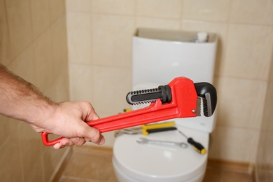 Photo of Plumber holding pipe wrench near toilet indoors, closeup