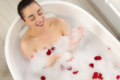 Happy woman taking bath in tub with foam and rose petals indoors, top view