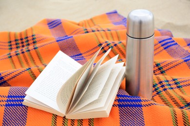 Photo of Metallic thermos with hot drink, open book and plaid on sandy beach. closeup