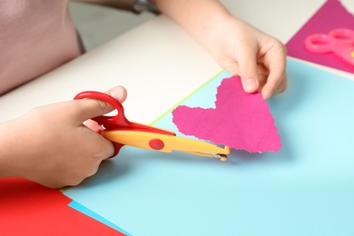 Photo of Child cutting out paper heart with craft scissors at table, closeup