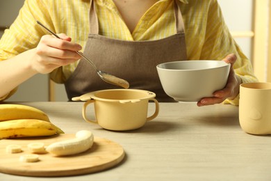 Photo of Healthy baby food. Woman putting delicious banana puree into bowl at light wooden table, closeup