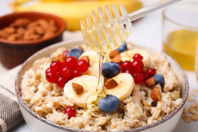 Honey pouring into bowl of oatmeal with berries, almonds and banana slices on table, closeup