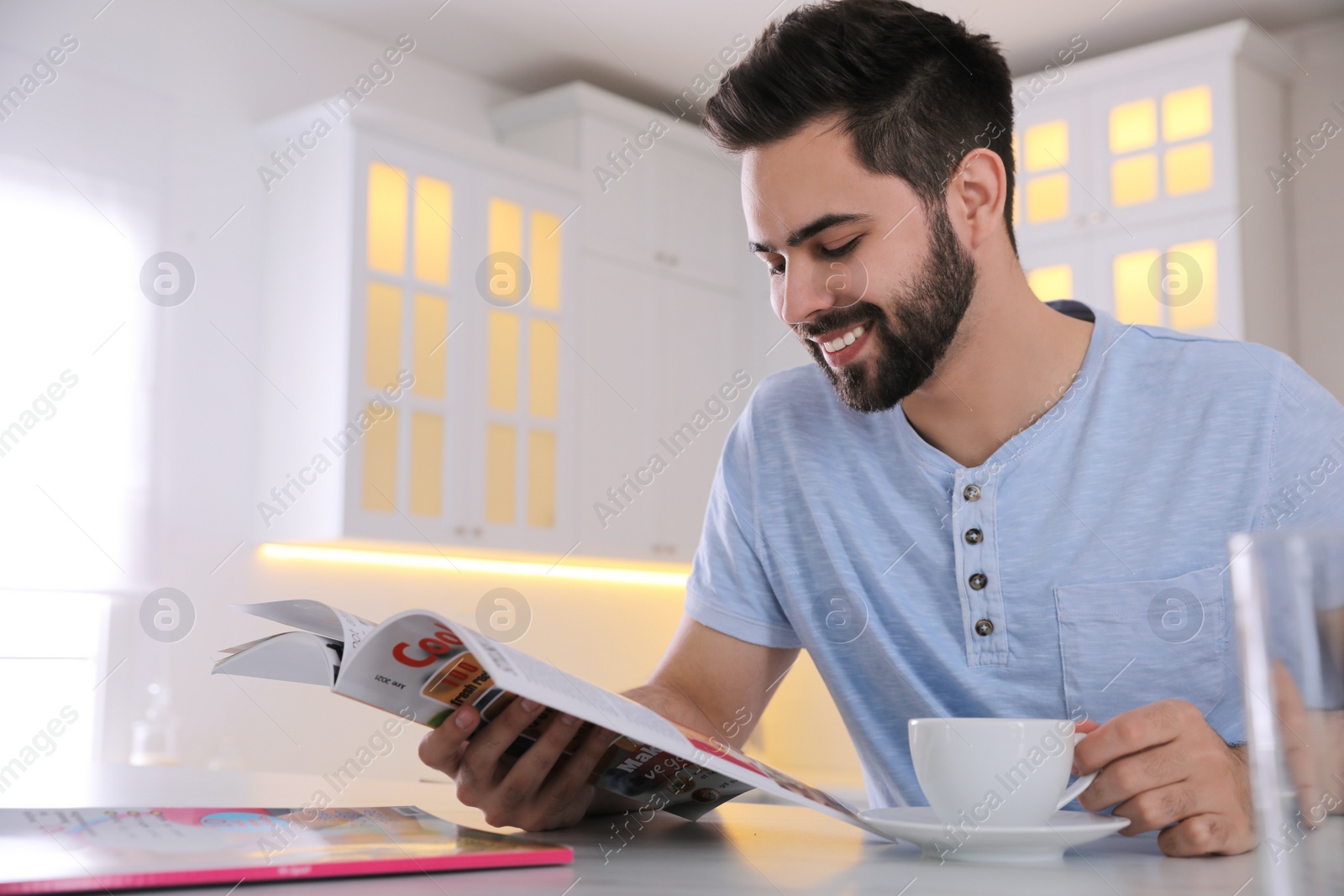 Photo of Young man with cup of drink reading magazine at table in kitchen