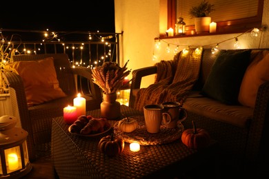 Rattan furniture, cups, fairy lights, burning candles and other autumn decor on outdoor terrace at night