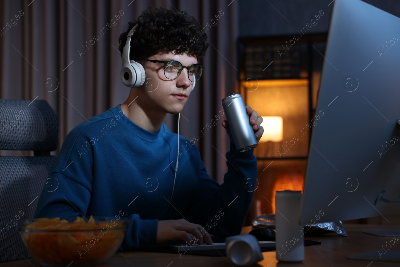 Photo of Young man with energy drink and headphones playing video game at wooden desk indoors