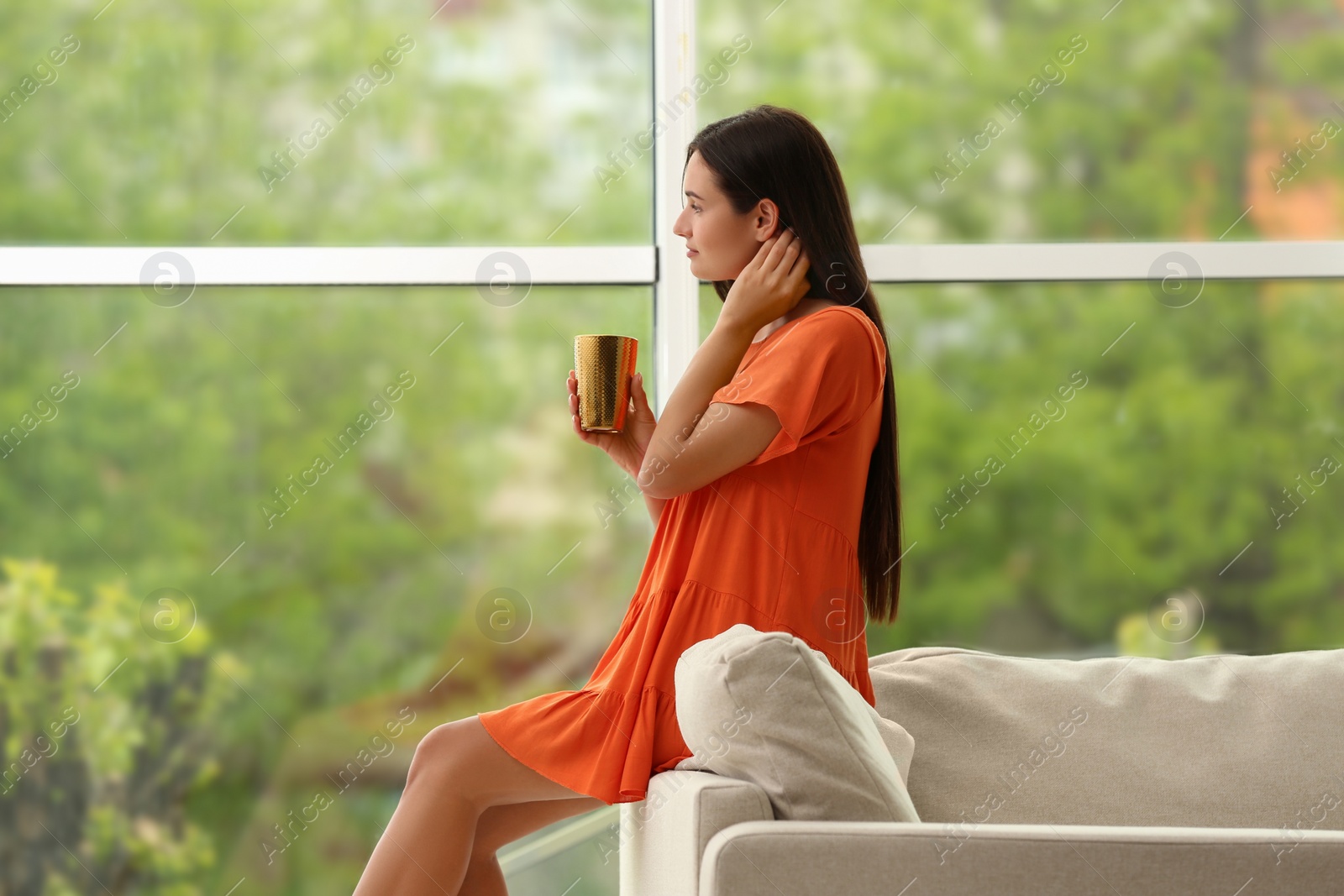 Photo of Young woman with cup of drink near sofa at home