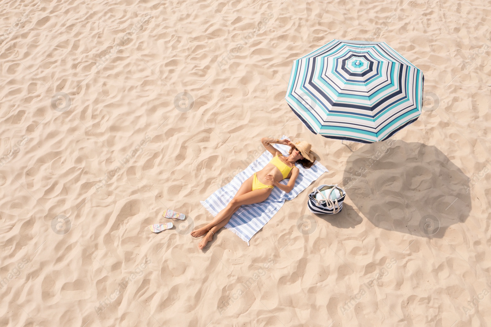 Image of Woman resting under striped beach umbrella at sandy coast