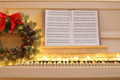 Photo of White piano with fairy lights, beautiful Christmas wreath and music sheets, closeup