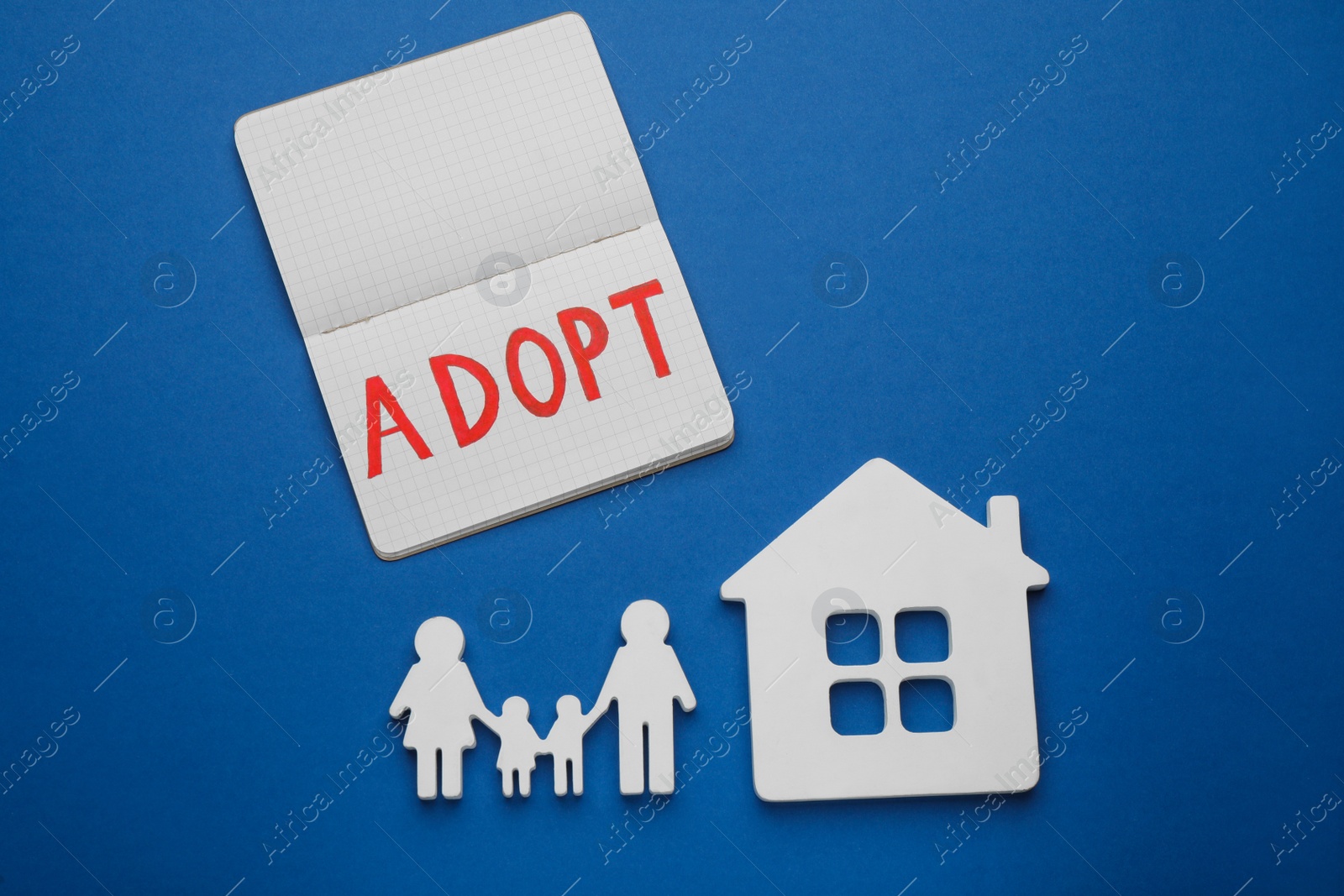 Photo of Family figure, house and notebook with word Adopt on blue background, flat lay