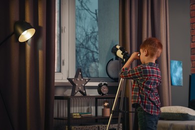 Photo of Little boy looking at stars through telescope in room, back view