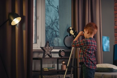 Little boy looking at stars through telescope in room, back view