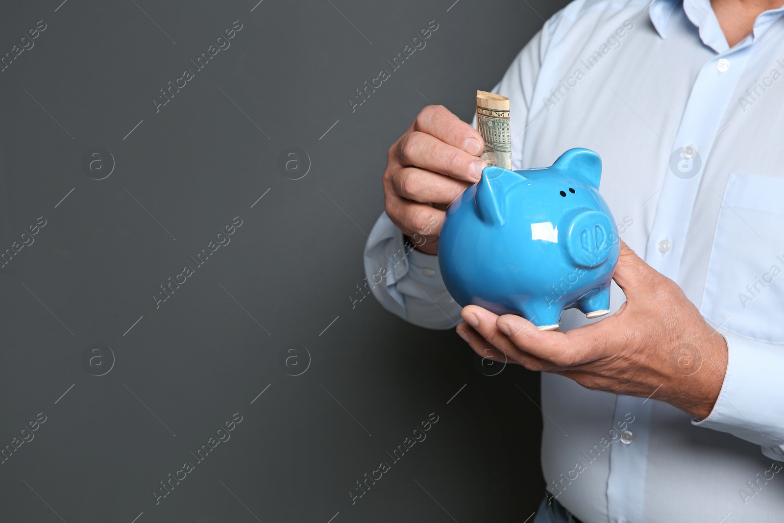 Photo of Mature man putting money into piggy bank on grey background, closeup. Space for text