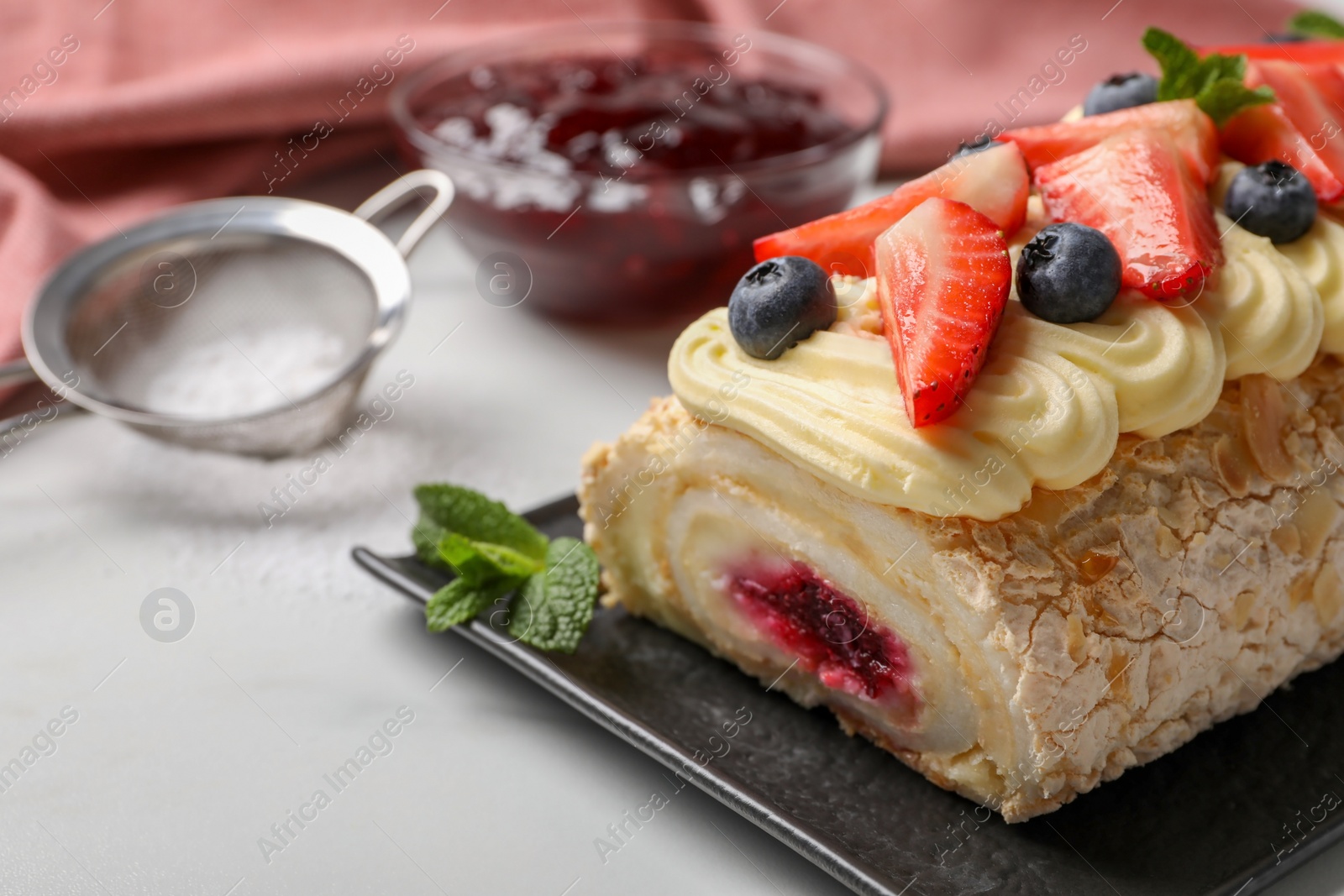 Photo of Tasty meringue roll with jam, cream, strawberry, blueberry and mint on white table, closeup. Space for text