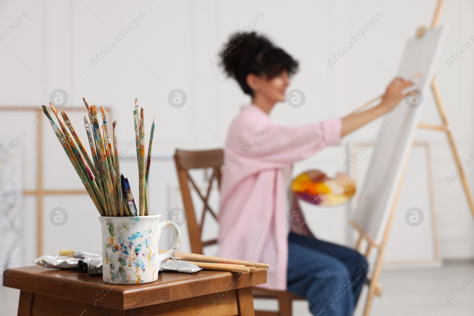 Photo of Young woman painting on easel with canvas in studio, focus on brushes