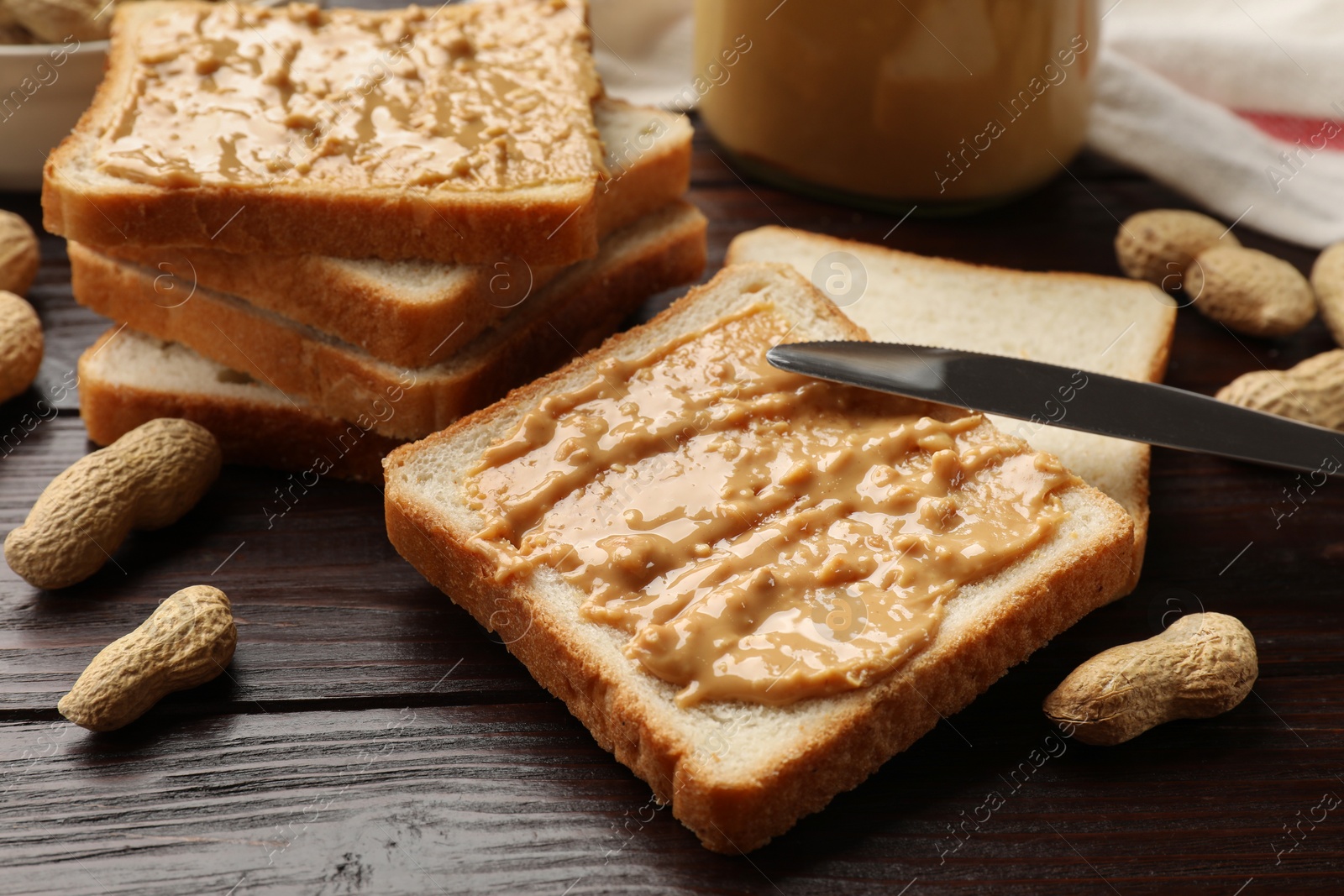 Photo of Delicious toasts with peanut butter, nuts and knife on dark wooden table, closeup