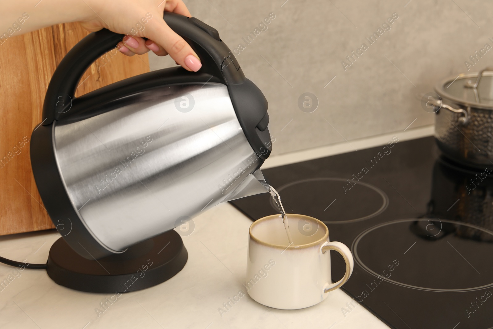 Photo of Woman pouring hot water from electric kettle into cup in kitchen, closeup