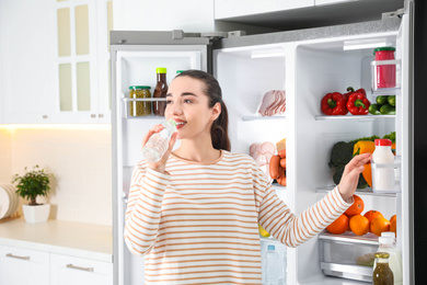 Photo of Young woman drinking water near open refrigerator in kitchen