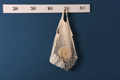 Conscious consumption. Net bag with eco friendly products hanging on blue wall indoors, space for text