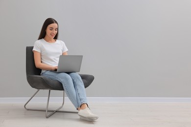 Beautiful woman using laptop while sitting in armchair near light grey wall indoors, space for text