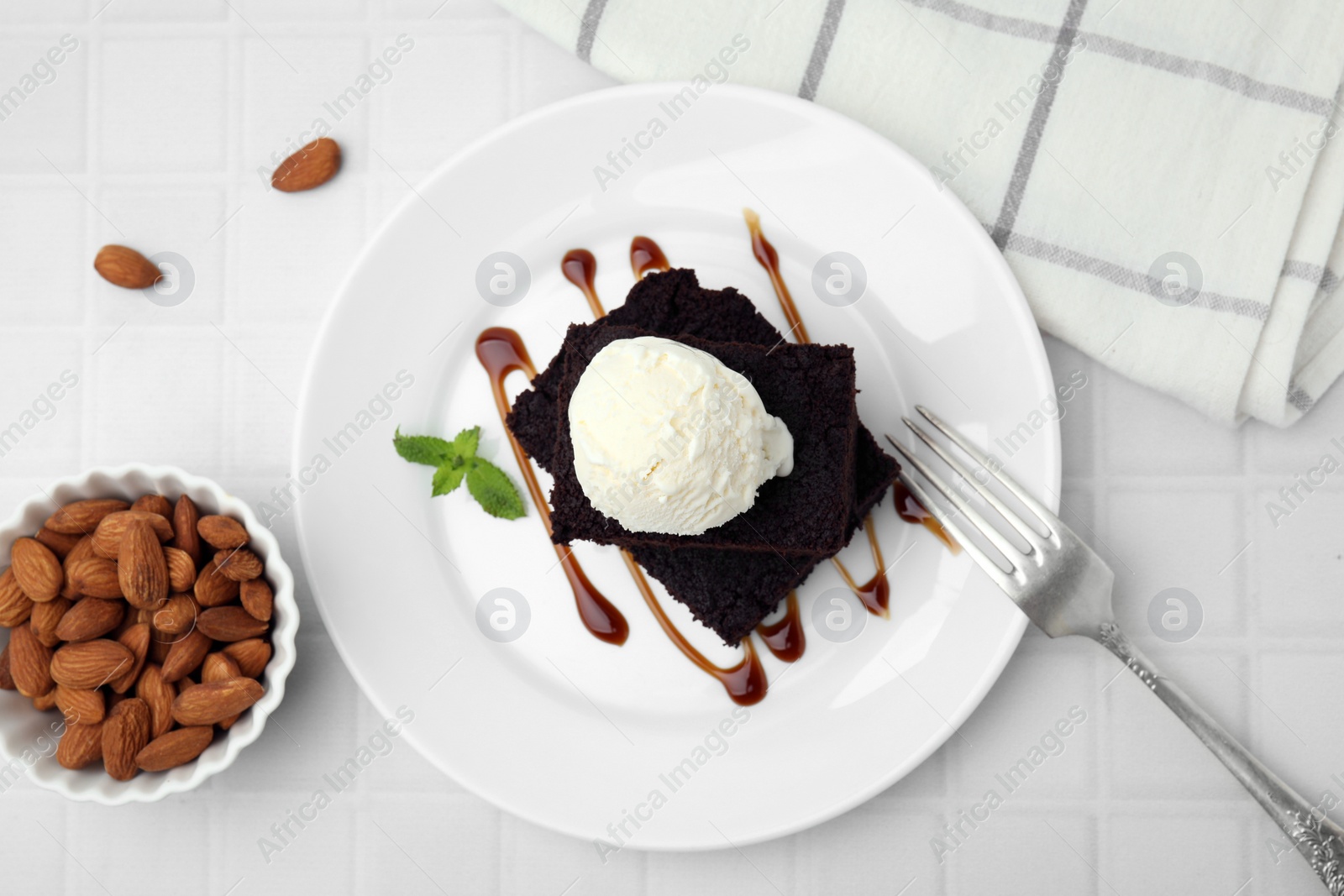 Photo of Tasty brownies served with ice cream and caramel sauce on white tiled table, flat lay