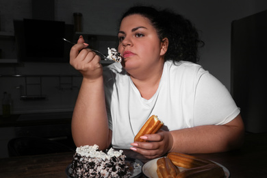 Photo of Depressed overweight woman eating sweets in kitchen at night