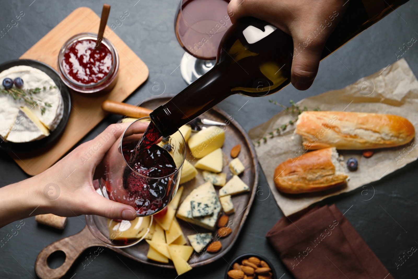 Photo of Man pouring red wine from bottle into glass over black table with snacks, top view