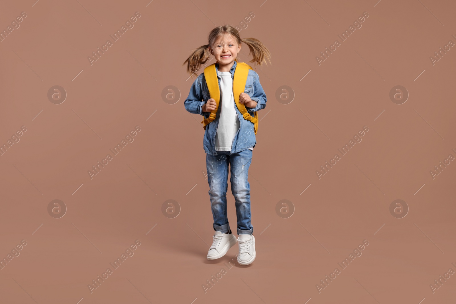 Photo of Happy schoolgirl with backpack jumping on brown background