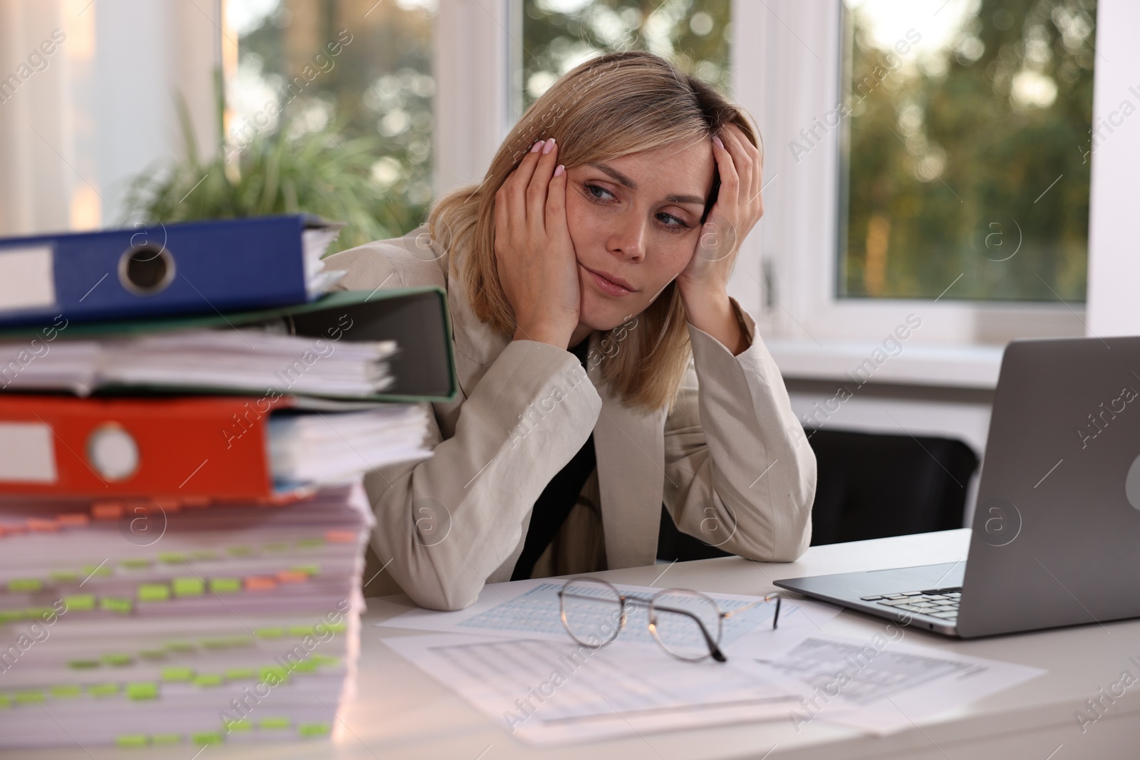 Photo of Overwhelmed woman sitting at table with stack of documents and folders in office